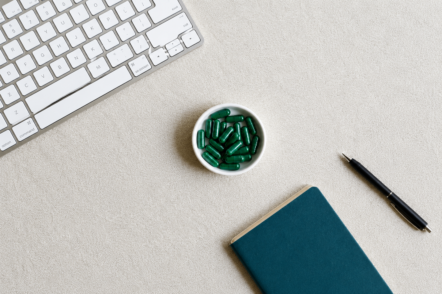 A small white bowl of green Synapt capsules on a light desk next to a keyboard, pen, and notebook.